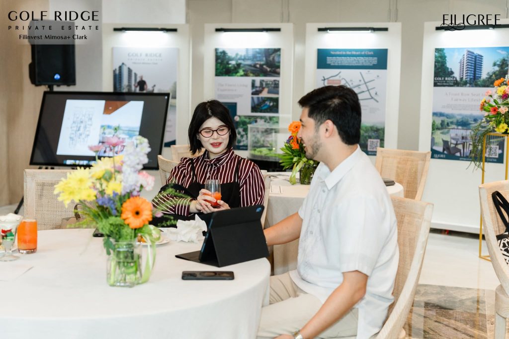 A couple sits at a table during an event at Golf Ridge Private Estate, with floral arrangements and displays of the properties in the background.