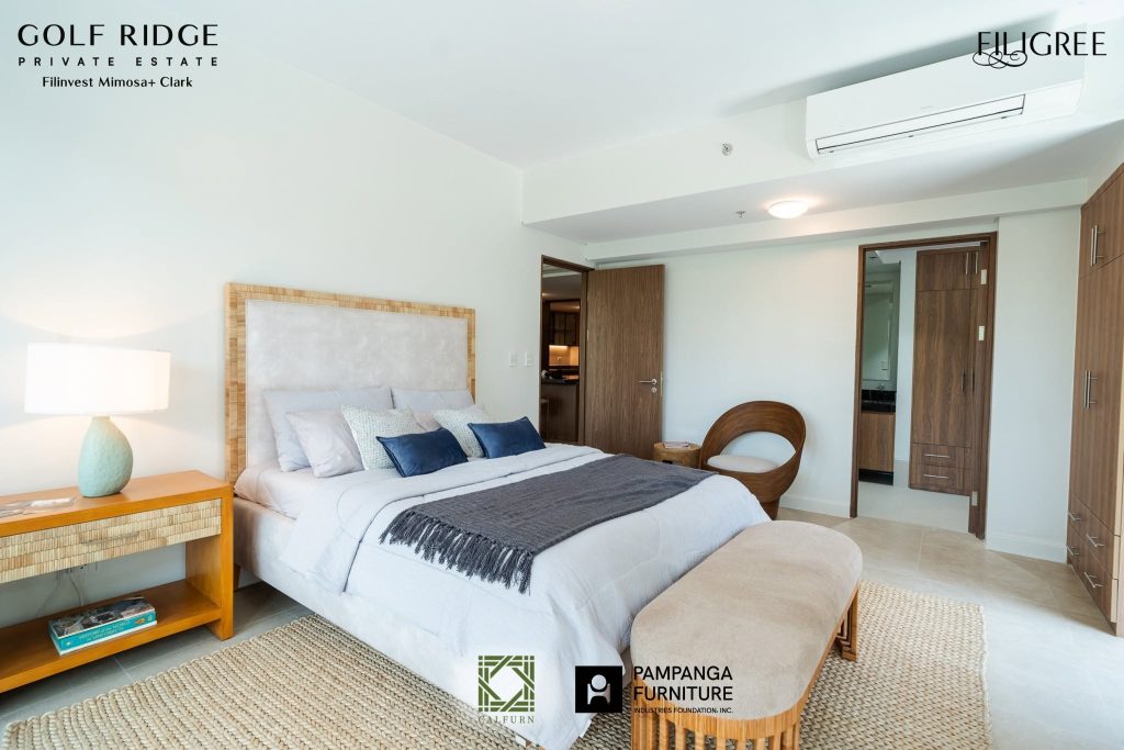 A modern, minimalist bedroom featuring a queen-sized bed with blue accent pillows, a woven headboard, wooden furniture, and a textured area rug.