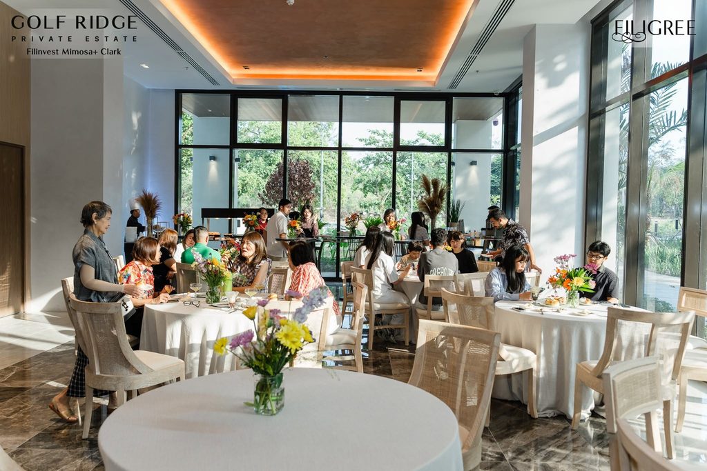 Guests dining at round tables decorated with fresh flowers inside a brightly banquet hall with large glass windows overlooking a garden.